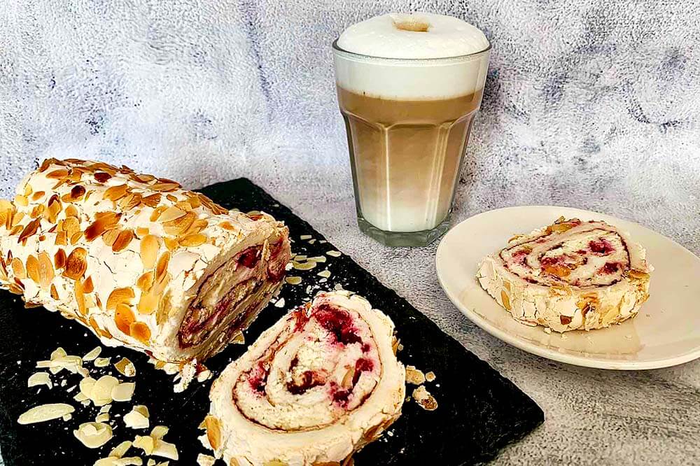 Pavlova roláda na bridlicovej tácke, plátok rolády na bielom tanieri a caffe latte v sklenenom pohári na sivom podklade