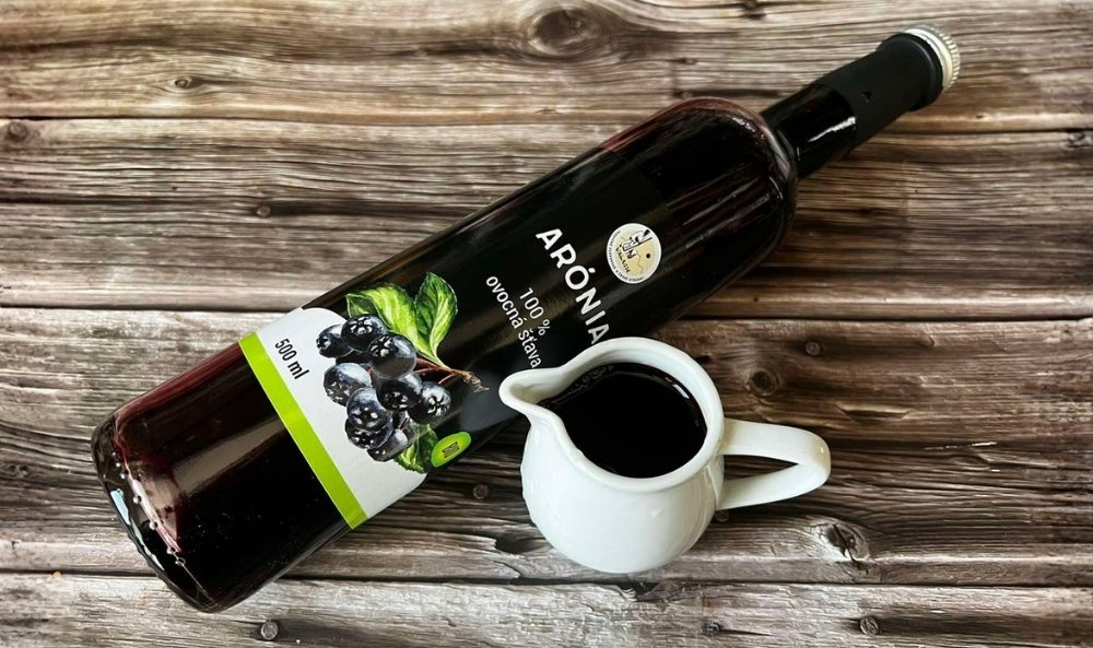 Fľaša aróniovej ovocnej šťavy a malý biely džbánik s nápojom na drevenom stole